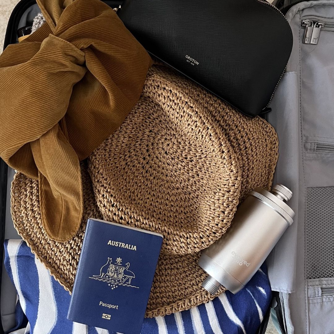 Evolved H2O shower filter packed in an open suitcase with a sun hat, passport and travel accessories