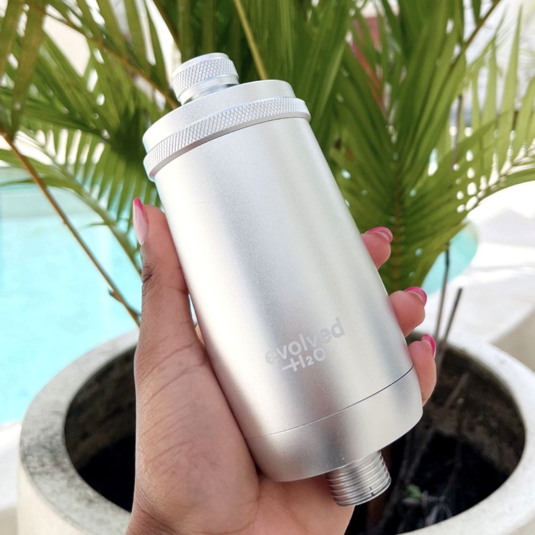 Silver Evolved H2O shower filter held in one hand beside a swimming pool and palm leaves