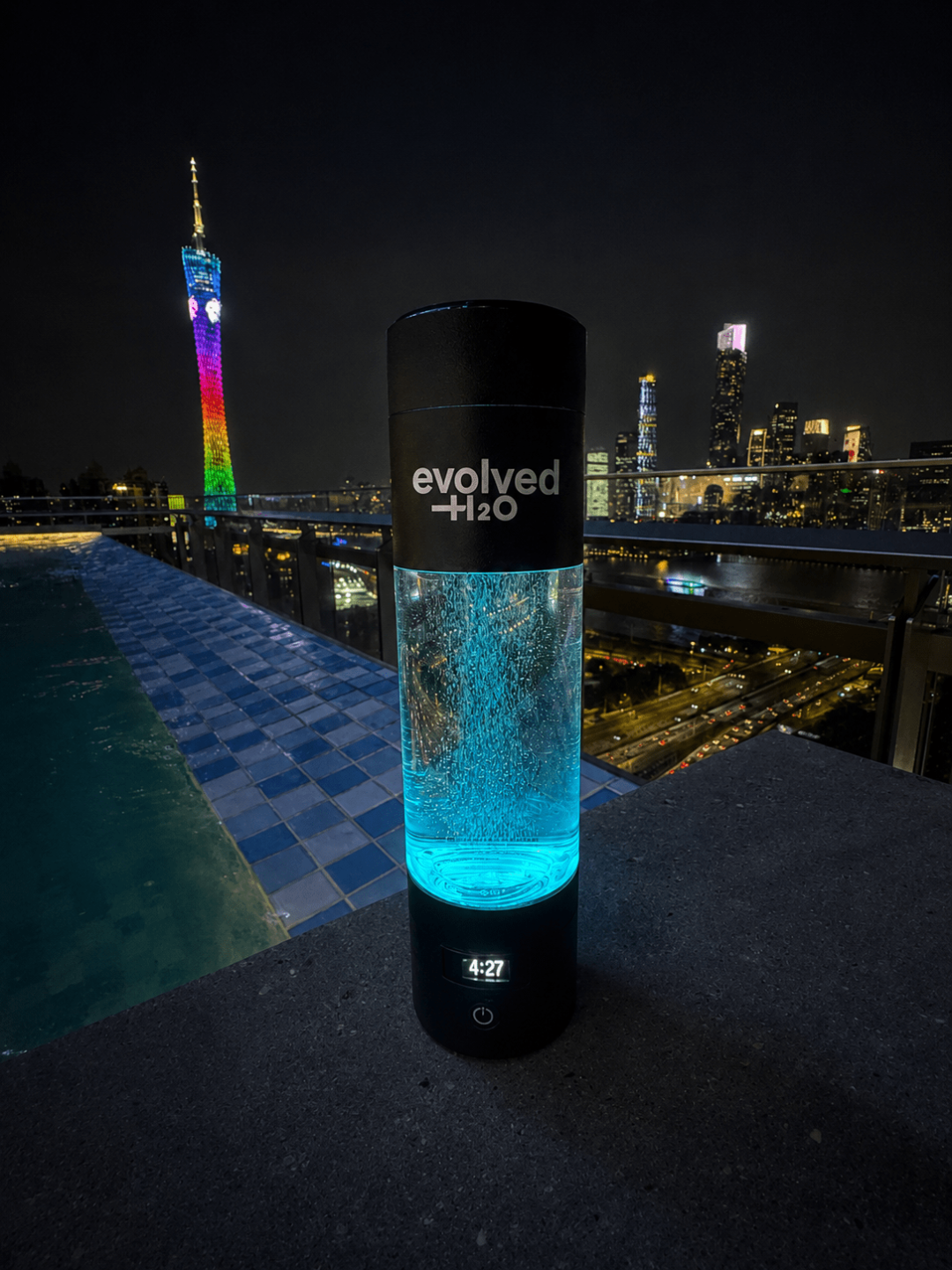 Evolved H2O Hydrogen Pro Bottle glowing blue beside a rooftop pool with a city skyline at night